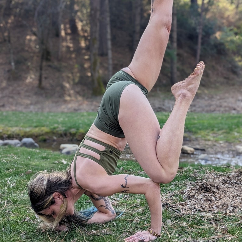 May include: A woman performs a yoga pose outdoors, wearing a green sports bra and matching shorts. She is balancing on her hands with one leg raised in the air.