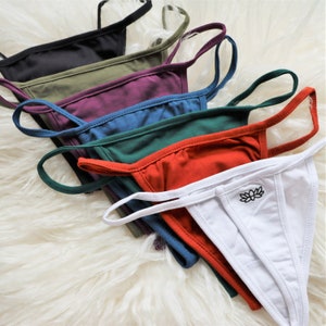 May include: A collection of thong underwear in various colors, including black, olive green, purple, blue, dark green, red-orange, and white. The white thong features a small black lotus flower emblem. The underwear is displayed on a fluffy white surface.