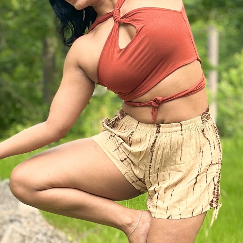 May include: A rust-colored halter top with a keyhole detail and wrap-around ties, paired with beige shorts featuring a brown striped pattern. The shorts have an elastic waistband and drawstring ties at the sides. The outfit is casual and summery.