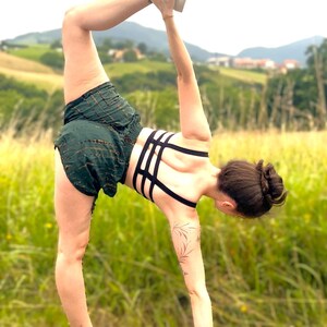 May include: A person in a yoga pose, wearing a dark green shorts and a black sports bra with a strappy design. The background is a field of tall grass and a distant view of rolling hills and trees.
