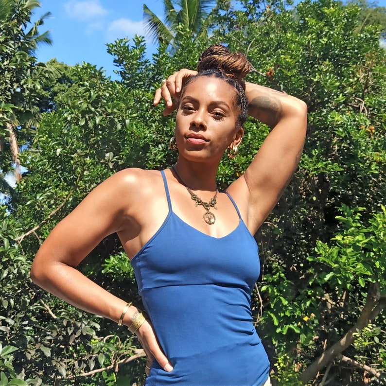 May include: A woman wearing a blue tank top with a gold necklace and earrings. She is standing in front of a lush green background.