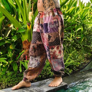 May include: Patchwork pants in shades of brown, pink, and purple. The loose-fitting pants feature a variety of patterned fabric squares, with an elastic waistband and gathered ankles. The pants are worn outdoors near lush greenery.