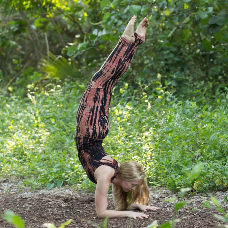 May include: A woman in a pink and black tie-dye jumpsuit performs a handstand in a forest setting.