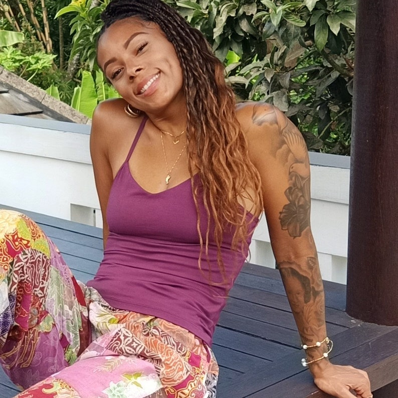 May include: A woman with long brown hair wearing a purple tank top and patterned pants. She is sitting on a wooden deck with a tattoo on her arm.