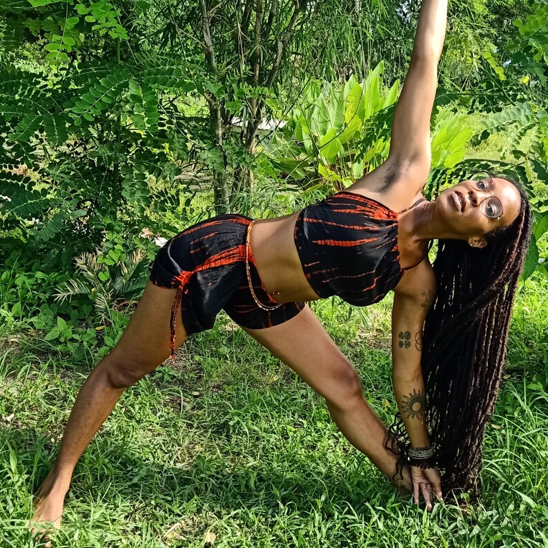 May include: A black and orange tie-dye yoga outfit, consisting of a crop top and shorts, is worn while doing a yoga pose. The outfit is accented with a gold chain. The person is wearing glasses and has long dreadlocks. The background is a lush green environment.