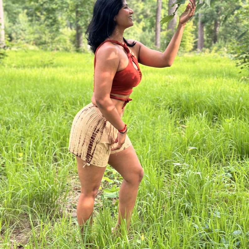 May include: A woman in a red halter top and beige shorts stands in a field of tall green grass. The shorts have a brown tie-dye pattern. The woman is reaching up towards a tree branch.