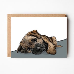 May include: A greeting card featuring a hand-drawn illustration of a Border Terrier puppy. The puppy is depicted in shades of brown and tan, with a black nose and large, expressive eyes. The card has a kraft paper border.