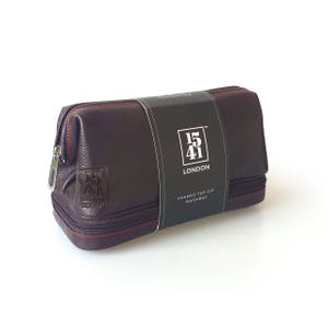 Peut inclure: Une trousse de toilette de voyage en cuir marron avec une fermeture à glissière. La trousse a une étiquette noire avec le texte "1541 LONDON" et "FRAMED TOP ZIP WASHBAG".