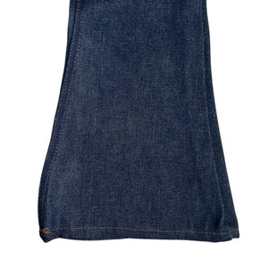 May include: Close-up of the bottom of a pair of dark blue denim bell bottom trousers. The trousers have a straight seam down the side and a hemmed bottom.
