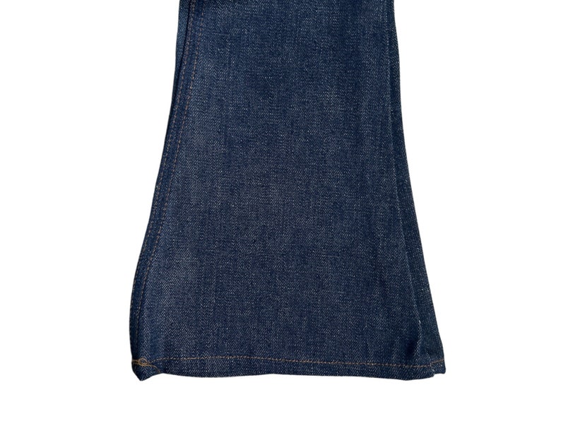 May include: Close-up of the bottom of a pair of dark blue denim bell bottom trousers. The trousers have a straight seam down the side and a hemmed bottom.
