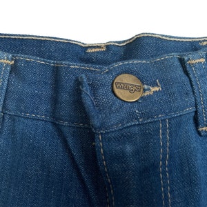 Może przedstawiać: Zbliżenie na pas pary niebieskich dżins&oacute;w z brązowym metalowym guzikiem z napisem "Wrangler".