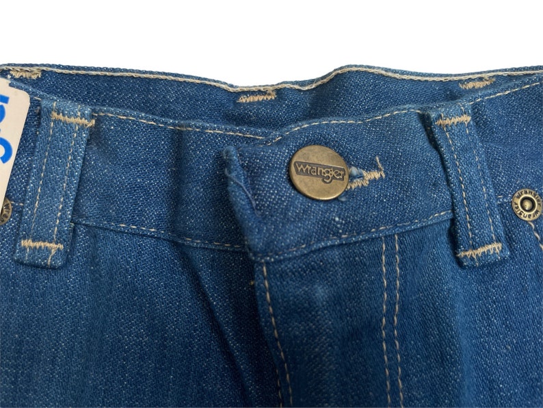 Może przedstawiać: Zbliżenie na pas pary niebieskich dżins&oacute;w z brązowym metalowym guzikiem z napisem "Wrangler".