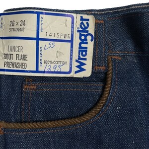 May include: A close-up of a pair of blue denim jeans with a brown corduroy waistband. The jeans have a label that reads "Wrangler" and "141SPWR". The label also includes the size "26 x 34", the model "Lancer Boot Flare Prewashed", and the content "100% Cotton".