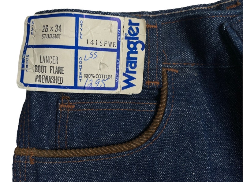 May include: A close-up of a pair of blue denim jeans with a brown corduroy waistband. The jeans have a label that reads "Wrangler" and "141SPWR". The label also includes the size "26 x 34", the model "Lancer Boot Flare Prewashed", and the content "100% Cotton".