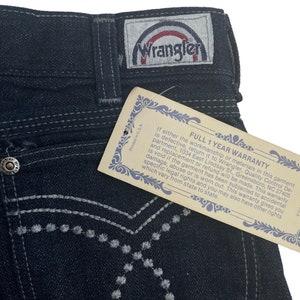 May include: A pair of dark blue denim jeans with a white embroidered design on the back pocket. The jeans have a Wrangler label with a rainbow logo. The tag attached to the jeans reads "Full 1 Year Warranty".