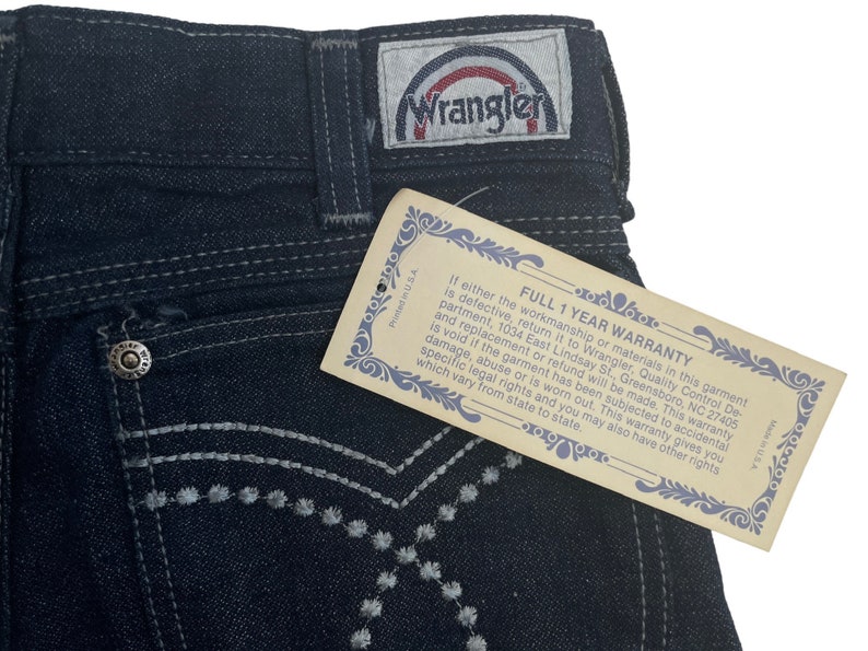May include: A pair of dark blue denim jeans with a white embroidered design on the back pocket. The jeans have a Wrangler label with a rainbow logo. The tag attached to the jeans reads "Full 1 Year Warranty".