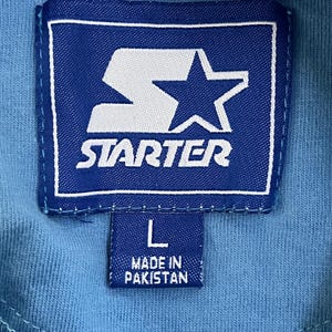 May include: A blue Starter brand tag with a white star and the word "STARTER" in white lettering. Below the tag is a smaller blue tag with the letter "L" and the text "MADE IN PAKISTAN".