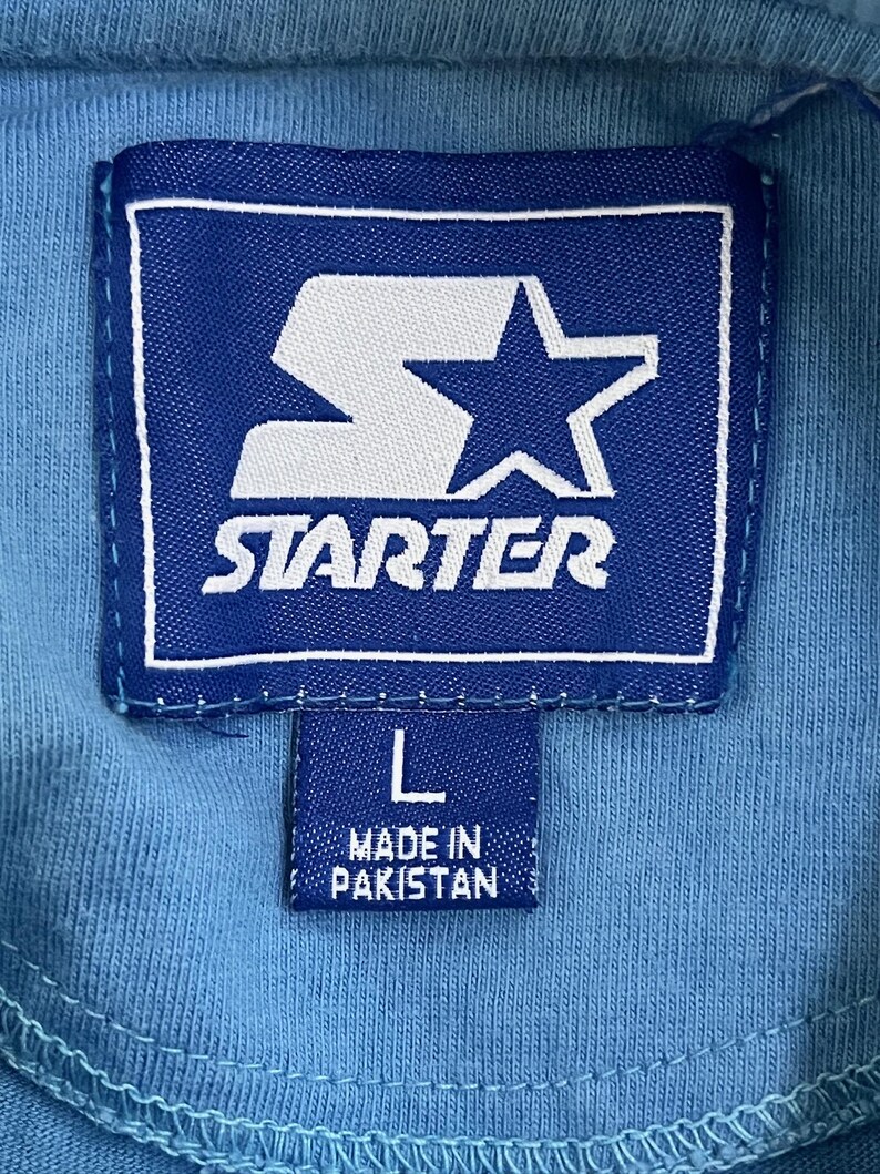 May include: A blue Starter brand tag with a white star and the word "STARTER" in white lettering. Below the tag is a smaller blue tag with the letter "L" and the text "MADE IN PAKISTAN".