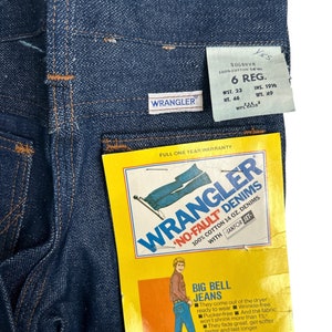 May include: A blue denim Wrangler 'No-Fault' jean tag with a full one year warranty. The tag features a man wearing bell bottom jeans and the text 'Big Bell Jeans'. The tag also includes the text '100% Cotton 14 oz. Denims with Sanfor-Set'.