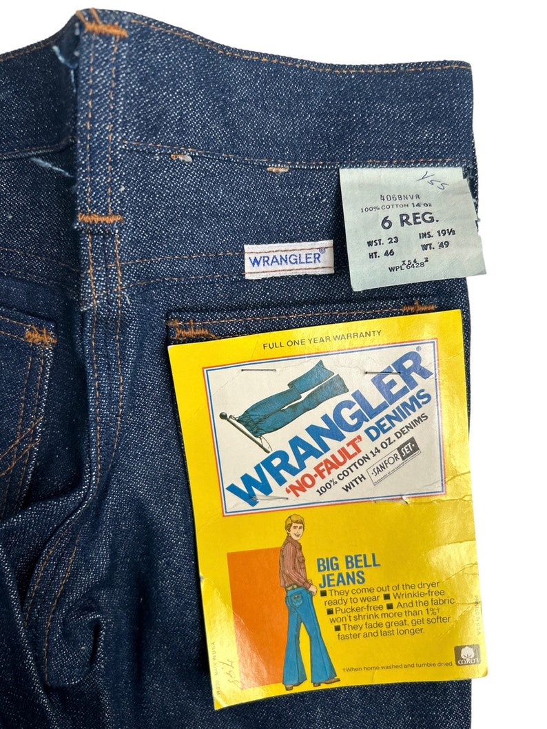 May include: A blue denim Wrangler 'No-Fault' jean tag with a full one year warranty. The tag features a man wearing bell bottom jeans and the text 'Big Bell Jeans'. The tag also includes the text '100% Cotton 14 oz. Denims with Sanfor-Set'.