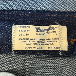 May include: A close-up of a blue denim jean label with white text. The label reads "STUDENT 537SPWR 26 X 30 Wrangler 'NO-FAULT' DENIMS R MACHINE WASH. TUMBLE DRY. AVOID OVER DRYING. REMOVE PROMPTLY. WASH DARKS SEPARATELY. MADE IN U.S.A. OHFFA35R"
