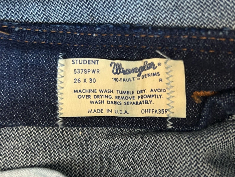 May include: A close-up of a blue denim jean label with white text. The label reads "STUDENT 537SPWR 26 X 30 Wrangler 'NO-FAULT' DENIMS R MACHINE WASH. TUMBLE DRY. AVOID OVER DRYING. REMOVE PROMPTLY. WASH DARKS SEPARATELY. MADE IN U.S.A. OHFFA35R"