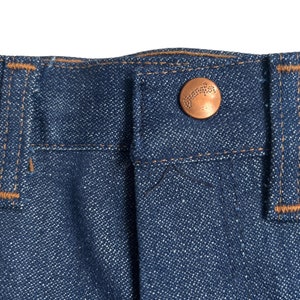 May include: Close-up of the waistband of a pair of blue denim jeans with a copper-coloured button that says "Wrangler" on it.