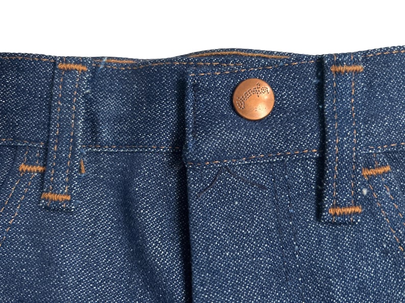 May include: Close-up of the waistband of a pair of blue denim jeans with a copper-coloured button that says "Wrangler" on it.