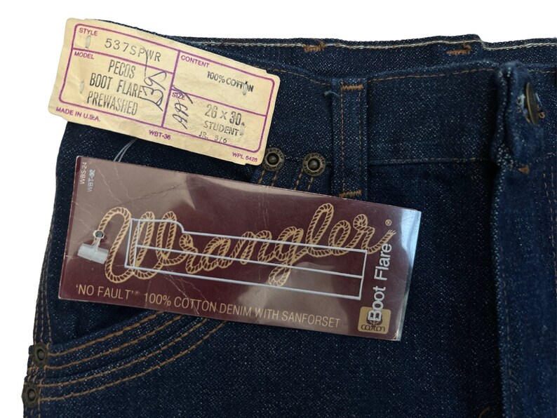 May include: A pair of blue denim jeans with a Wrangler label. The jeans are a boot cut style and have a pre-washed finish. The label says "No Fault" 100% cotton denim with Sanforset. The size tag says 26 x 30, Student.