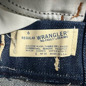 May include: A white tag with black text attached to a pair of blue denim jeans. The tag reads "6 Regular Wrangler 'No-Fault' Denims" and includes care instructions: "Machine wash, tumble dry, avoid overdrying, remove promptly. Wash darks separately. E NF-20 Made in U.S.A."