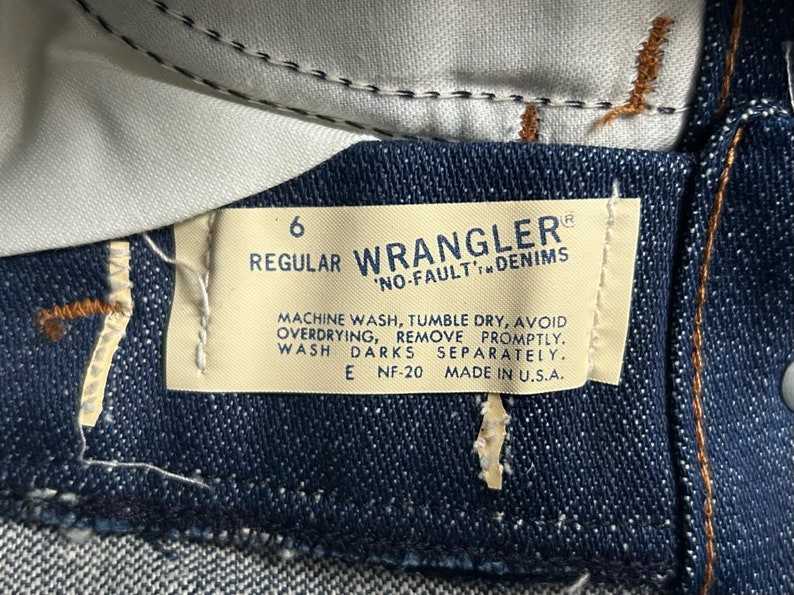May include: A white tag with black text attached to a pair of blue denim jeans. The tag reads "6 Regular Wrangler 'No-Fault' Denims" and includes care instructions: "Machine wash, tumble dry, avoid overdrying, remove promptly. Wash darks separately. E NF-20 Made in U.S.A."