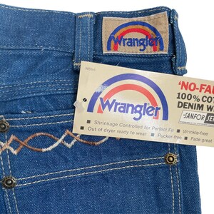 Może przedstawiać: Dżinsy z niebieskiego denimu z haftowanym wzorem w kolorze brązowym i białym na tylnej kieszeni. Dżinsy mają metkę Wrangler z logo tęczy i tekstem 'NO-FAULT' 100% COTTON DENIM WITH SANFOR SET'.