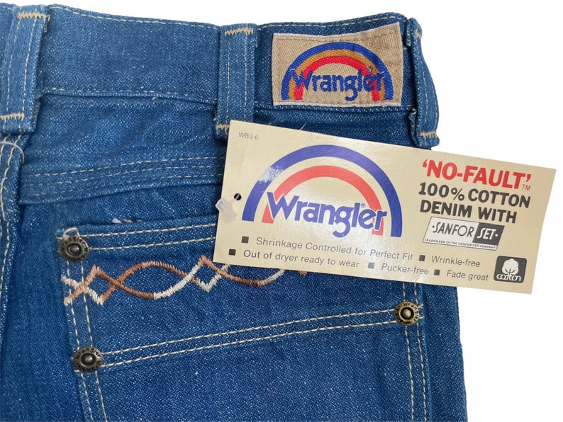 Może przedstawiać: Dżinsy z niebieskiego denimu z haftowanym wzorem w kolorze brązowym i białym na tylnej kieszeni. Dżinsy mają metkę Wrangler z logo tęczy i tekstem 'NO-FAULT' 100% COTTON DENIM WITH SANFOR SET'.