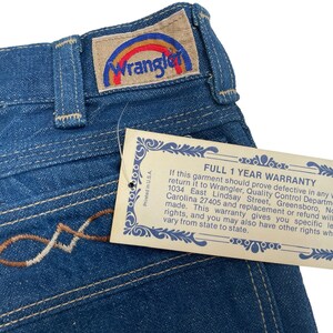 Może przedstawiać: Dżinsy z niebieskiego denimu z haftowanym wzorem w kolorze brązowym i białym na kieszeni. Dżinsy mają metkę Wrangler z logo tęczy. Do dżins&oacute;w dołączona jest metka gwarancyjna z tekstem "FULL 1 YEAR WARRANTY" i szczeg&oacute;łami dotyczącymi gwarancji.