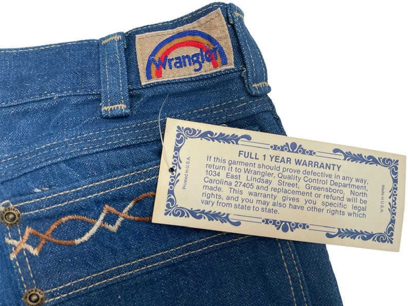 Może przedstawiać: Dżinsy z niebieskiego denimu z haftowanym wzorem w kolorze brązowym i białym na kieszeni. Dżinsy mają metkę Wrangler z logo tęczy. Do dżins&oacute;w dołączona jest metka gwarancyjna z tekstem "FULL 1 YEAR WARRANTY" i szczeg&oacute;łami dotyczącymi gwarancji.