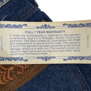 May include: A close-up of a blue denim jean with a brown leather patch and a yellow warranty tag. The tag reads "Full 1 Year Warranty" and includes details about the warranty and contact information for Wrangler.