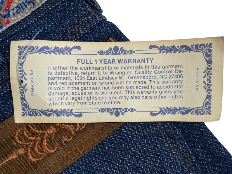 May include: A close-up of a blue denim jean with a brown leather patch and a yellow warranty tag. The tag reads "Full 1 Year Warranty" and includes details about the warranty and contact information for Wrangler.