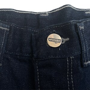 May include: Close-up of the waistband of a pair of dark blue denim jeans with a silver Wrangler button.