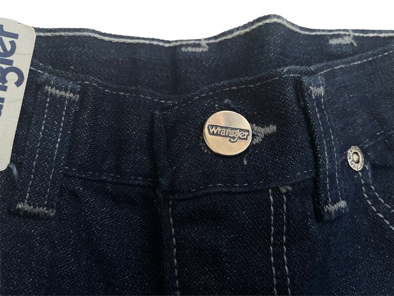 May include: Close-up of the waistband of a pair of dark blue denim jeans with a silver Wrangler button.