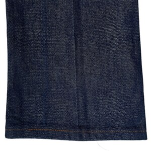 May include: A close-up of the bottom hem of a pair of dark blue denim jeans. The hem is sewn with a brown thread.