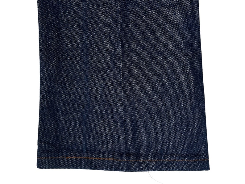 May include: A close-up of the bottom hem of a pair of dark blue denim jeans. The hem is sewn with a brown thread.