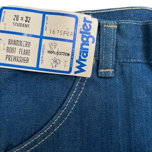 Może przedstawiać: Etykieta dżins&oacute;w Wrangler z niebieskiego denimu z białymi szwami. Etykieta zawiera napis "Wrangler" białymi literami i białą metkę z następującymi informacjami: "Rozmiar 26 x 32 Student", "Model Bandolero Boot Flare Prewashed", "Styl 167SFBR", "Skład 100% bawełna".