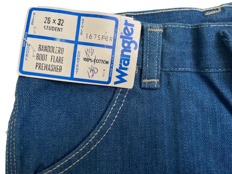 Może przedstawiać: Etykieta dżins&oacute;w Wrangler z niebieskiego denimu z białymi szwami. Etykieta zawiera napis "Wrangler" białymi literami i białą metkę z następującymi informacjami: "Rozmiar 26 x 32 Student", "Model Bandolero Boot Flare Prewashed", "Styl 167SFBR", "Skład 100% bawełna".