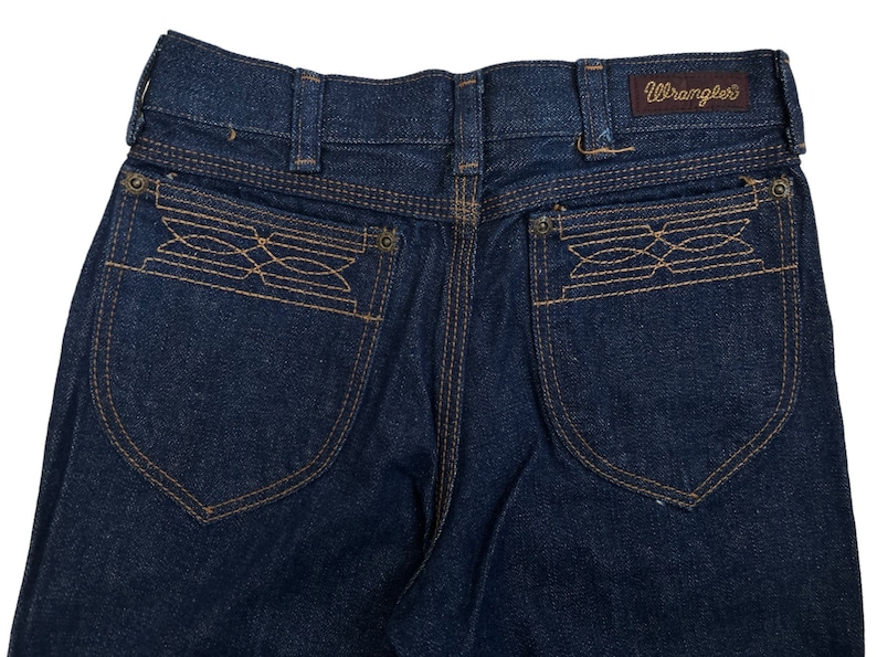 May include: A pair of dark blue denim jeans with a Wrangler label on the back waistband. The back pockets have a unique embroidered design.