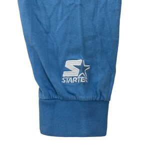 May include: Close-up of the blue cuff of a long-sleeved shirt with a white embroidered "STARTER" logo and a star.