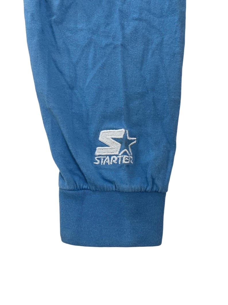 May include: Close-up of the blue cuff of a long-sleeved shirt with a white embroidered "STARTER" logo and a star.