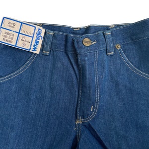 Może przedstawiać: Para niebieskich dżins&oacute;w z jasnobrązowymi szwami. Dżinsy mają metkę z napisem "Wrangler" i "26 x 32" z tekstem "BANDOLERO BOOT FLARE PREWASHED".