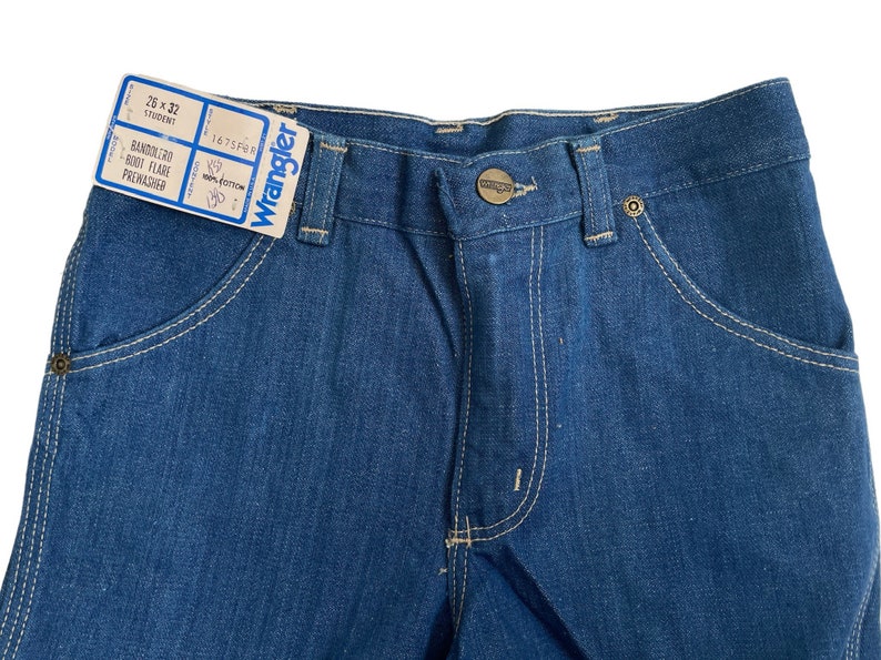 Może przedstawiać: Para niebieskich dżins&oacute;w z jasnobrązowymi szwami. Dżinsy mają metkę z napisem "Wrangler" i "26 x 32" z tekstem "BANDOLERO BOOT FLARE PREWASHED".