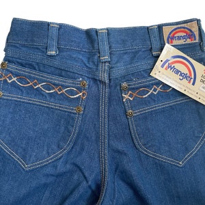 Może przedstawiać: Dżinsy z niebieskiego denimu z haftowanym wzorem w kolorze brązowym i białym na tylnych kieszeniach. Dżinsy mają metkę Wrangler i metkę z napisem "No-Fault 100% Cotton Denim with Sanfor Set."