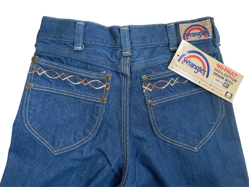Może przedstawiać: Dżinsy z niebieskiego denimu z haftowanym wzorem w kolorze brązowym i białym na tylnych kieszeniach. Dżinsy mają metkę Wrangler i metkę z napisem "No-Fault 100% Cotton Denim with Sanfor Set."